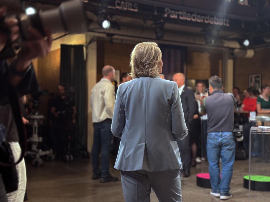 Sylvi Listhaug (Frp) st&aring;r med ryggen til kamera under TV 2s partilederdebatt i Oslo. Hun har p&aring; seg en lys buksedress. Rundt henne er fotografer, TV-team og tilskuere samlet.