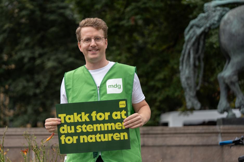 En mann if&oslash;rt gr&oslash;nn vest med MDG logo p&aring;, st&aring;r med ett skilt hvor det st&aring;r "Takk for at du stemmer for naturen"
