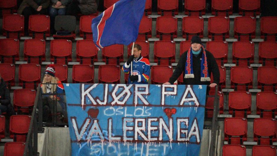 En supporter veiver med et flagg fremfor en banner med teksten "Kjør på Vålerenga" 