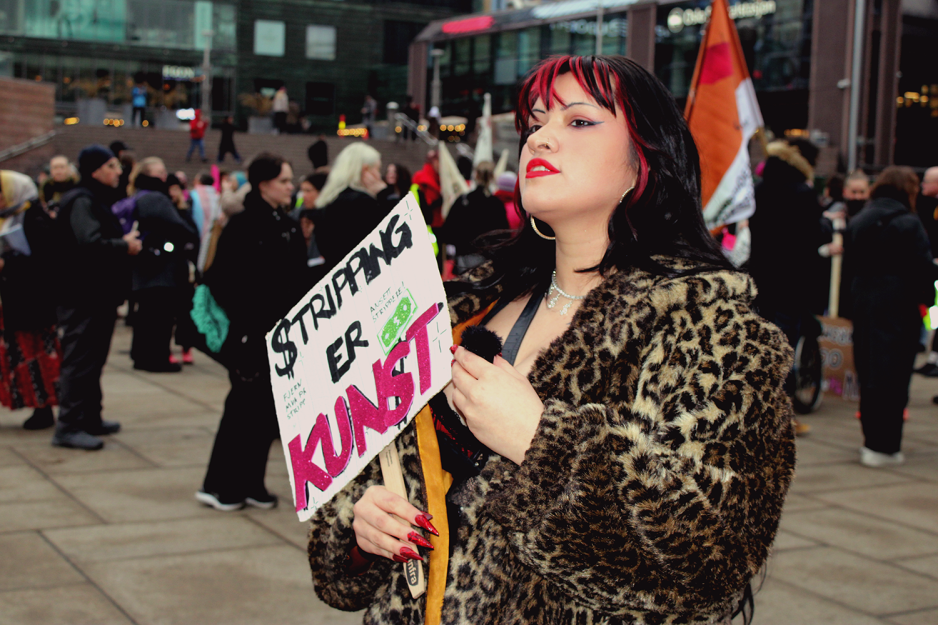 Stripper Alma Adoreholder en plakat der det står "Stripping er Kunst". Med haka rettet oppover ser hun bestemt ut