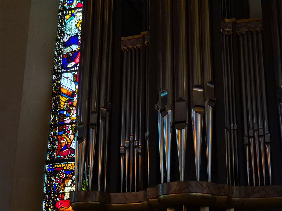 N&aelig;rbilde av orgelpiper ved siden av et langt glassvindu i Oslo domkirke. M&oslash;rk stemning, men det kommer sterke, livlige farger fra vinduet.