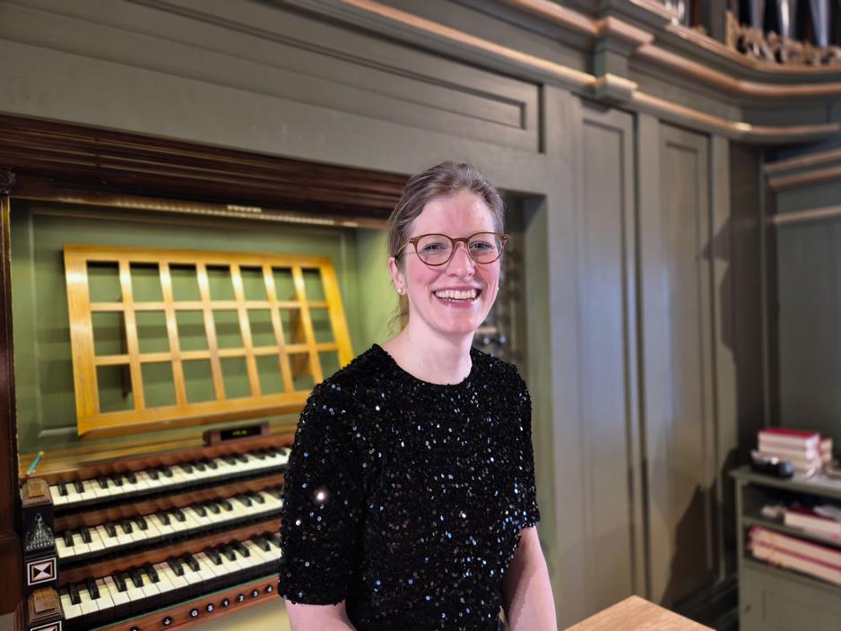 En svartkledd dame sitter foran et orgel. Hun har satt opp h&aring;ret og har p&aring; et par briller. Hun smiler mot kamera.