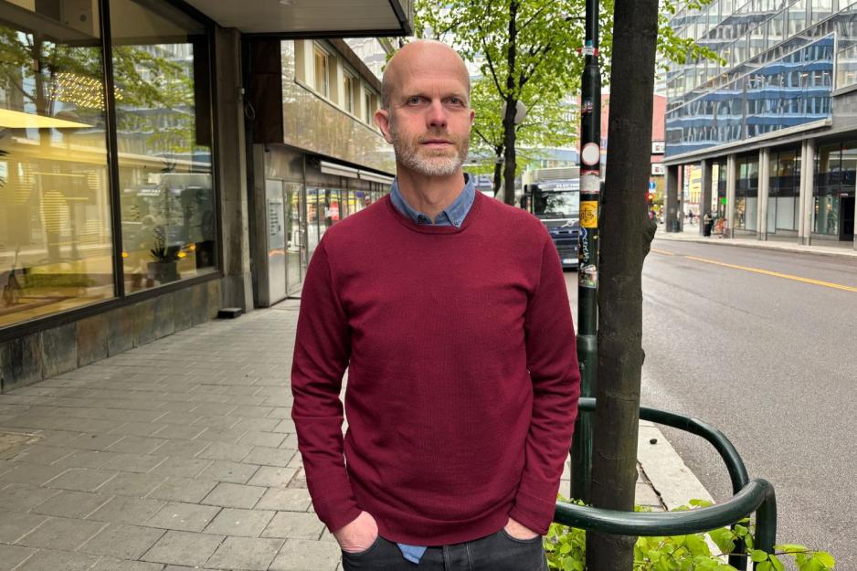 Mann med r&oslash;d genser st&aring;r med hendene i lomma ute i gata. 