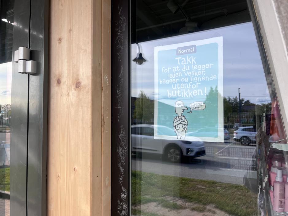 På bildet ser vi en plakat som har blitt hengt opp utenfor Normalbutikken. På plakaten står det "Takk for at du legger igjen vesker, bagger og lignende utenfor butikken!"
