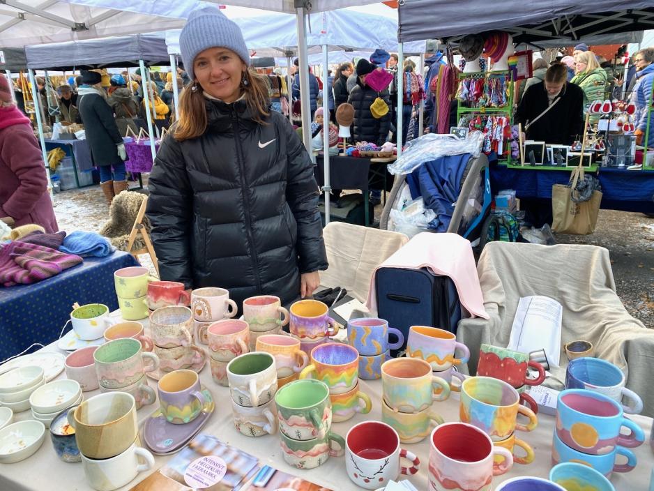 Bildet viser en kvinne som smiler til kameraet. Hun st&aring;r overfor flere keramikk-krus av ulike farger og motiver.