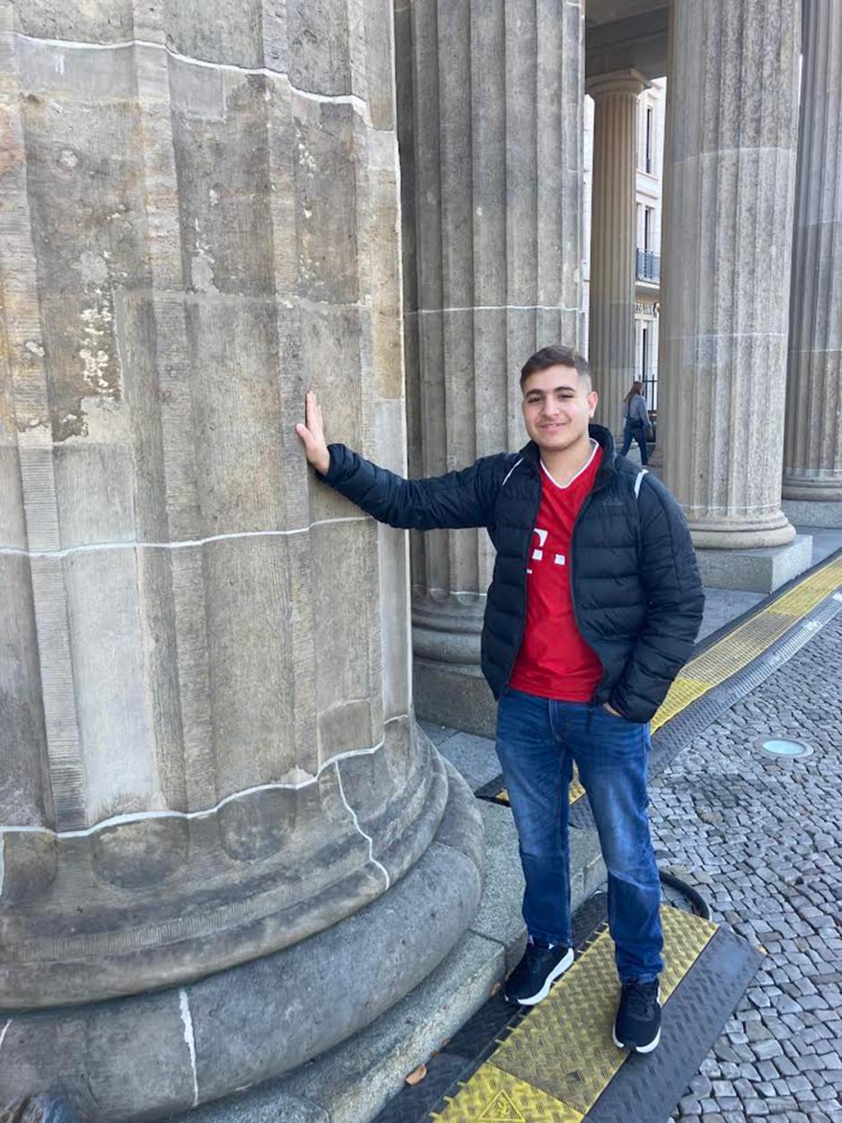 ung gutt som lener seg intill en s&oslash;yle med armen. gutter har p&aring; m&oslash;rkebl&aring; olabukse, r&oslash;d genser og en m&oslash;rkebl&aring; jakke over 