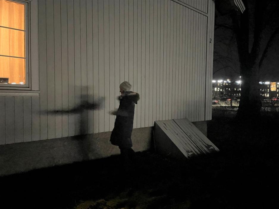 En dame st&aring;r foran en hvit husvegg og veiver med armene. Det er m&oslash;rkt, og skyggen hennes vises p&aring; husveggen. Damen har p&aring; seg vinterkl&aelig;r. 