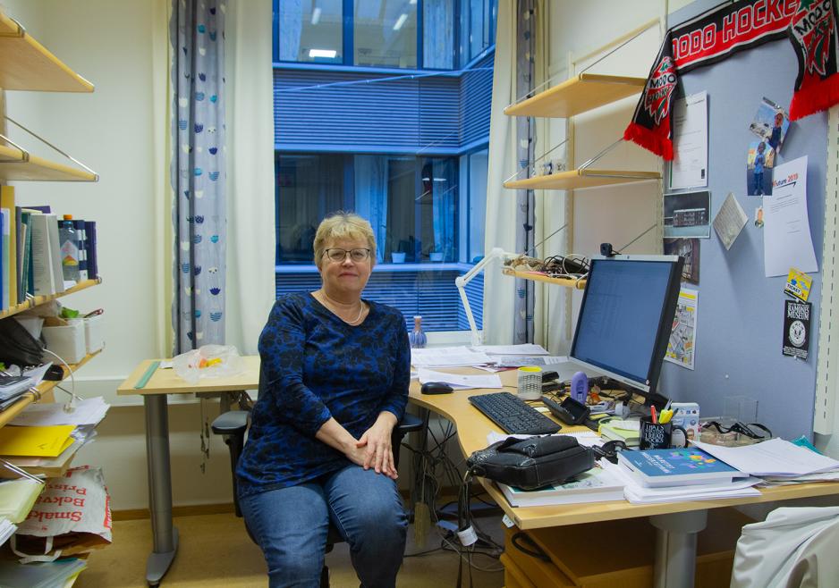 En dame med lyst h&aring;r og briller sitter p&aring; kontoret sitt, med en PC og en rotete pult bak seg. 