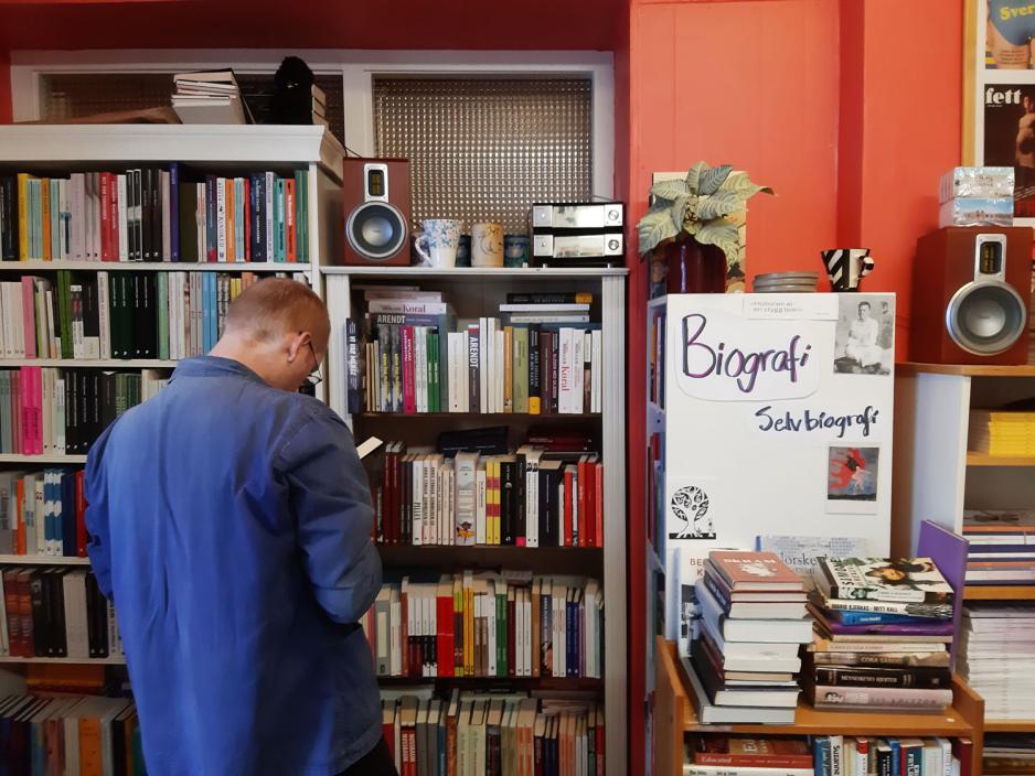 En bokhylle. En mann st&aring;r vendt mot bokhyllen med ryggen mot kameraet.