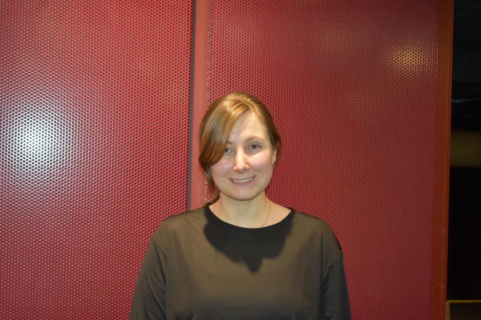 Portrett av ei blid dame med brunt oppsatt h&aring;r. Hun st&aring;r foran en r&oslash;d vegg. 