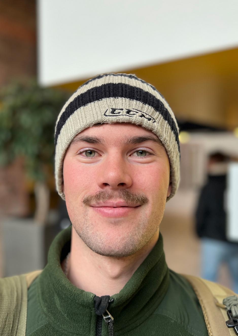 Mann med brun bart, lue p&aring; hodet og et mildt smil