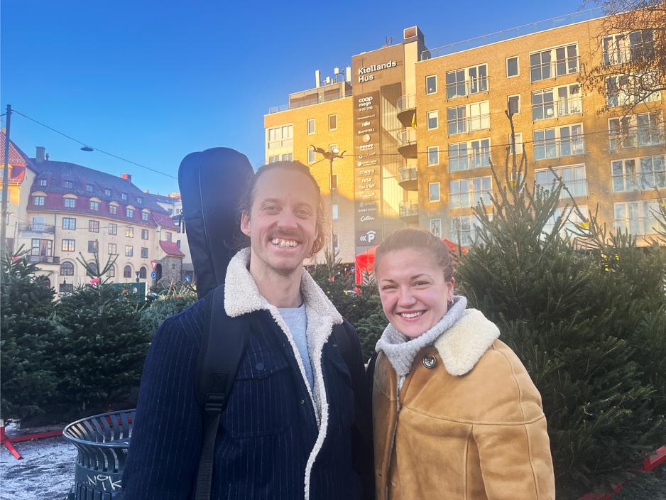 En mann med m&oslash;rk bl&aring; jakke og en gitar p&aring; ryggen og en kvinne med lys brun skinnjakke med pels i kragen. 