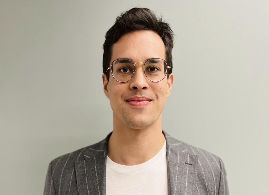 Daniel Myklebust smiler foran en hvit vegg, if&oslash;rt gr&aring; dressjakke, briller og hvit t-skjorte. 