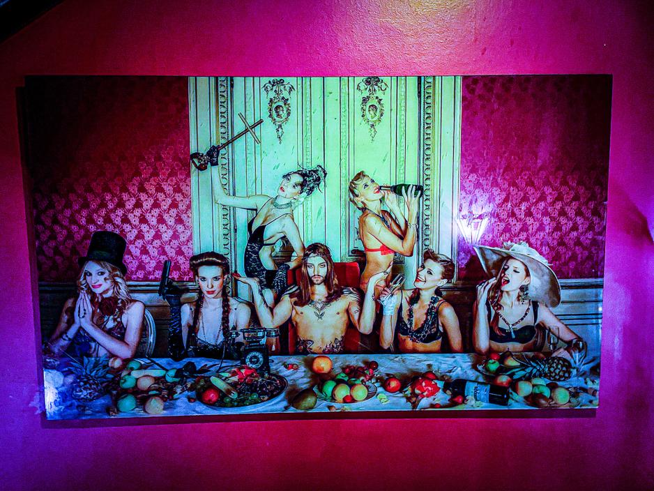 Et bilde henger p&aring; en r&oslash;d vegg. Bildet er av lettkledde mennesker som spiser middag i undert&oslash;yet. En har en pistol, en annen har et kors. 