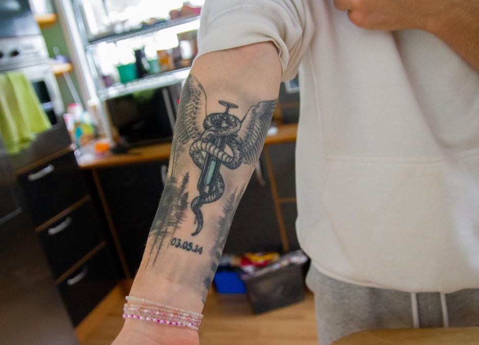 Tatovering p&aring; underarm, av en spr&oslash;yte med en slange rundt. P&aring; toppen er det vinger. Under spr&oslash;yten er det en dato. 