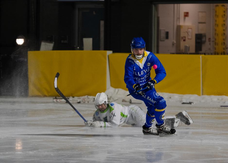 Bandyspiller sl&aring;r ballen mens motstanderen ligger p&aring; isen bak