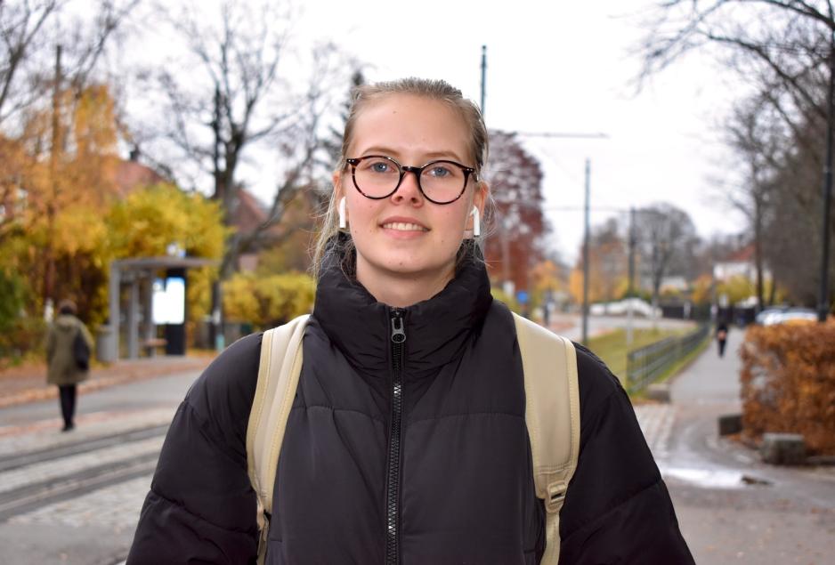 Ung kvinne med m&oslash;rke briller, blondt h&aring;r, svart jakke og beige ryggsekk venter p&aring; trikken. Hun smiler. 