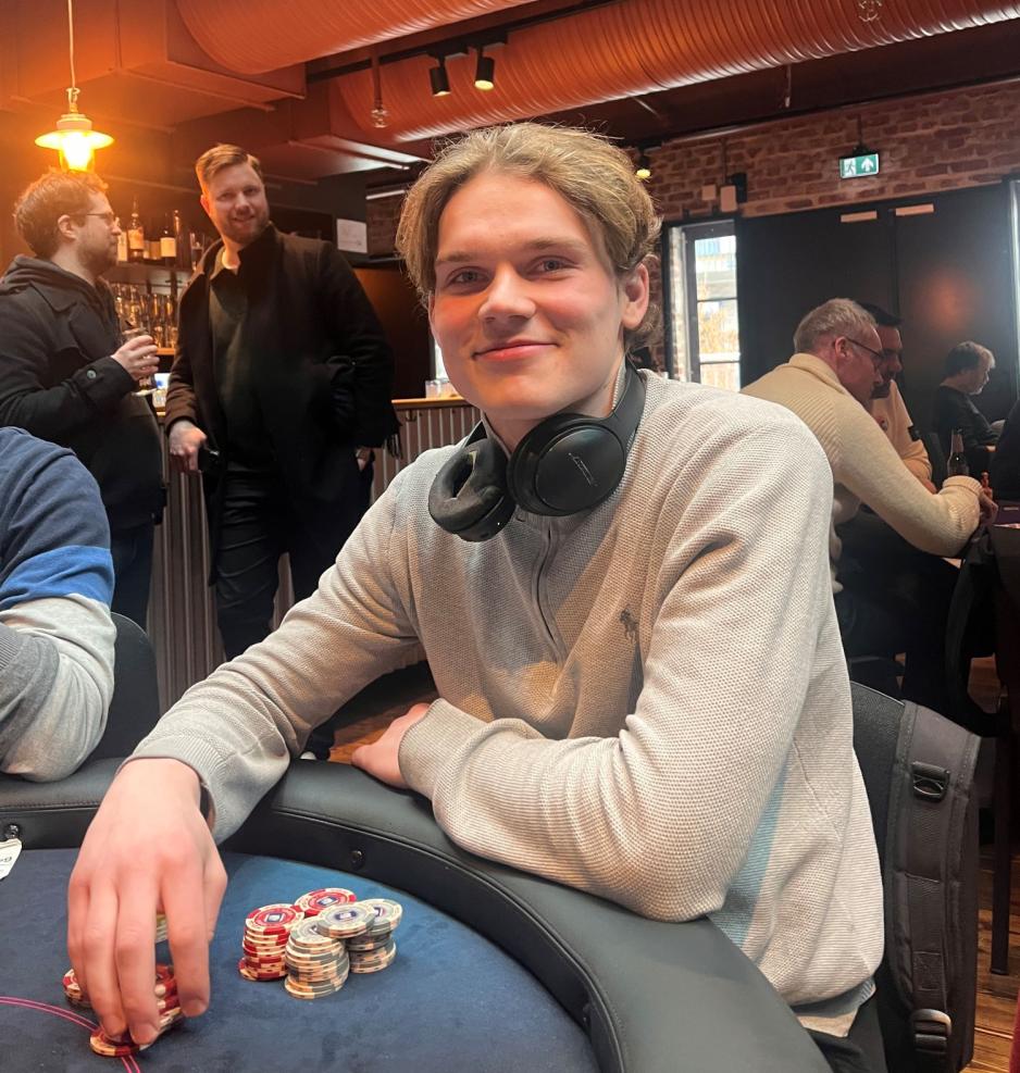 Smilene ung mann i gr&aring; genser og hodetelefoner rundt nakken, sitter ved et bord og spiller poker.
