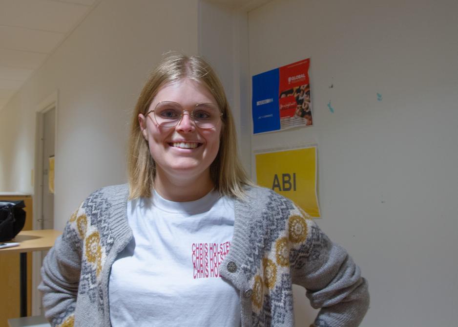 En ung jente med briller smiler mot kamera. Hun har lyst h&aring;r, og har p&aring; seg en hvit t-skjorte med en m&oslash;nstret strikkejakke over. 