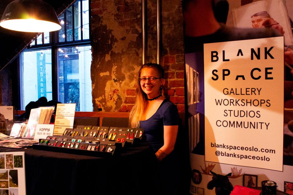 Dame som sitter bak et bord med mange &oslash;redobber og noen postkort. til h&oslash;yre for henne er en stor plakat der det st&aring;r blank space-logoen. 