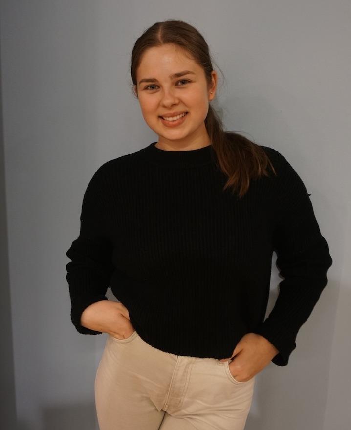 Jente med brunt h&aring;r poserer inntil en bl&aring;lig vegg. Jenta smiler vennlig inn i kameraet. 