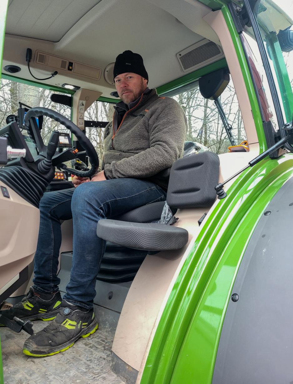 Mann i gr&aring; jakke i f&oslash;rersetet p&aring; en gr&oslash;nn traktor.