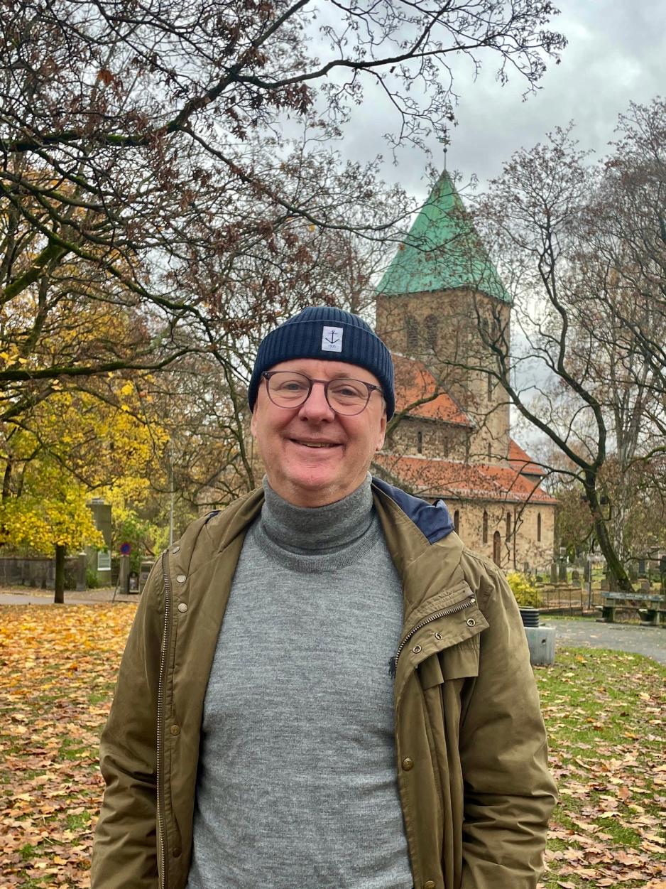 Bildet viser Finn Folke, som er kommunikasjonsansvarlig i Det kirkelige fellesr&aring;det i Oslo. Han har p&aring; seg briller og lue, og er avbildet foran Gamle Aker kirke p&aring; St Hanshaugen i Oslo. 