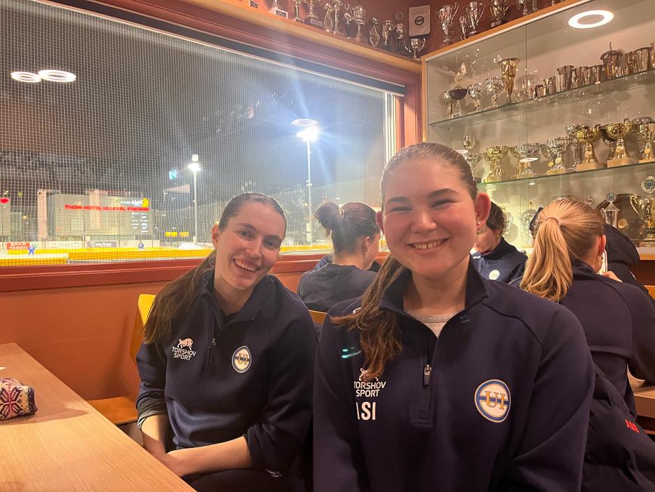 To kvinner sitter ved et bord og smiler til kamera. De har begge p&aring; m&oslash;rk bl&aring; treningsgenser