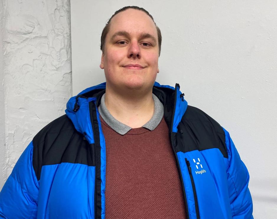 En mann ved en hvit vegg som har p&aring; seg ei bl&aring; jakke. 