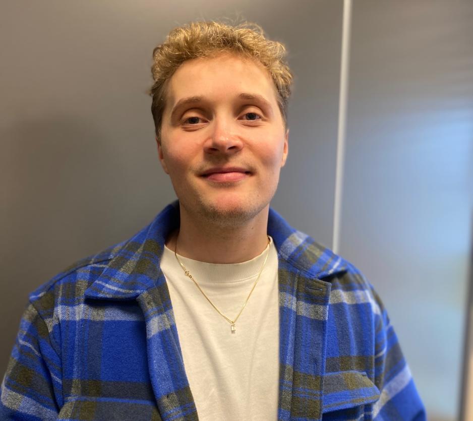 Bildet viser et portrettbilde av en mann med blondt, kr&oslash;llete h&aring;r som smiler til kameraet. Han har p&aring; seg bl&aring;, rutete jakke.