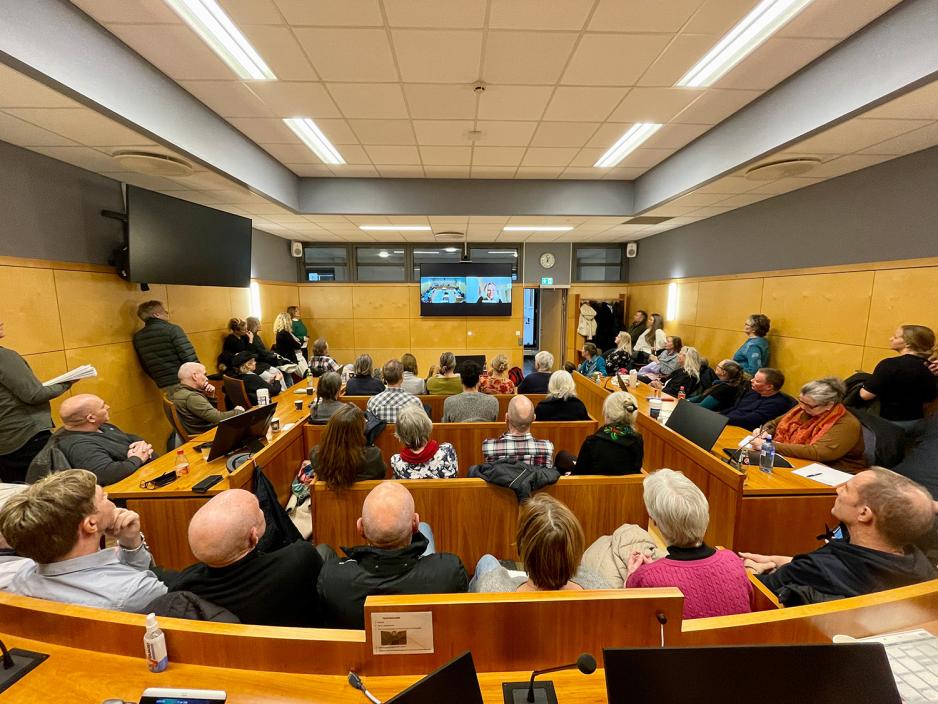 P&aring; bildet ser man bakhodet til mange mennesker. De ser p&aring; en skjerm. De sitter i en sal med brune trepanelvegger. De lytter. 