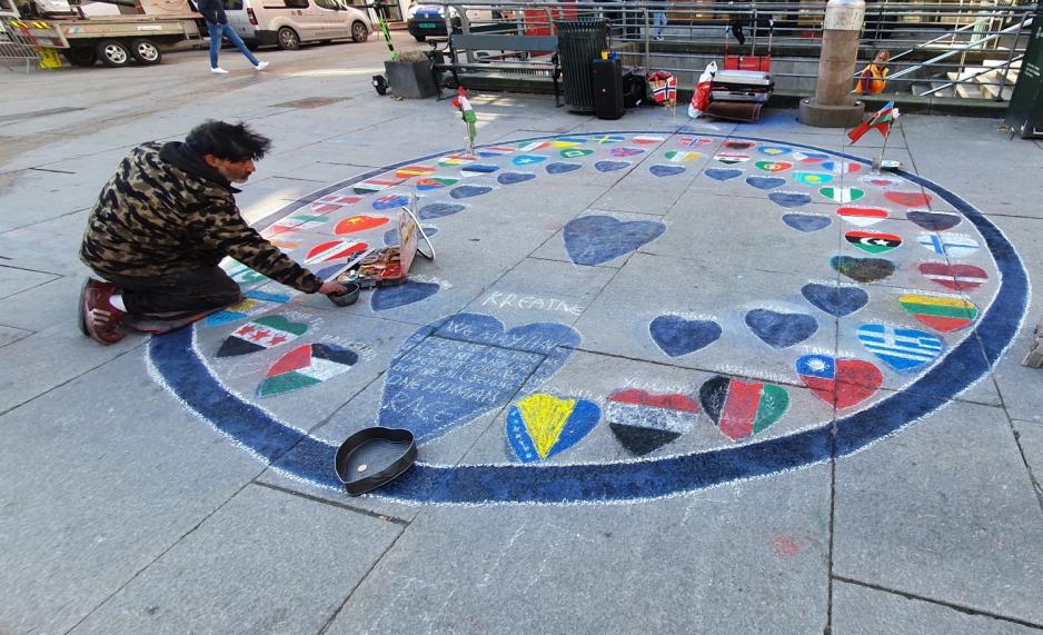 Bilde av en mann som sitter p&aring; kn&aelig;rne. P&aring; bakken foran han er det en sirkel med diverse flagg. 