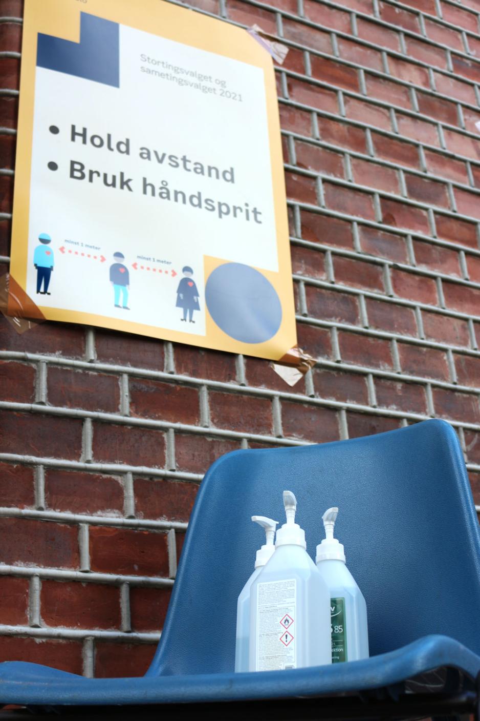 Bilde av tre spritflaskar p&aring; ein stol foran eit skilt om smittevern. 