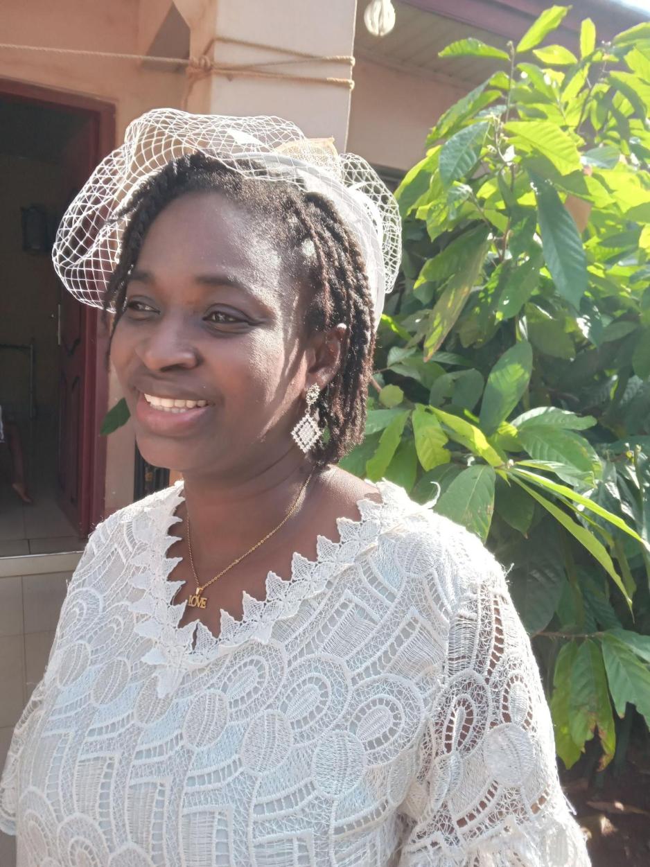 En kvinne i hvit kjole smiler med blikket vendt fra kamera. Bak henne er det gr&oslash;nne planter.