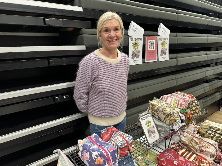 En smilende dame som st&aring;r bak et bord fylt med h&aring;ndlagde toalettmapper