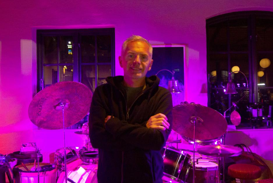 En mann med svart jakke st&aring;r med armene foldet foran to trommesett. 