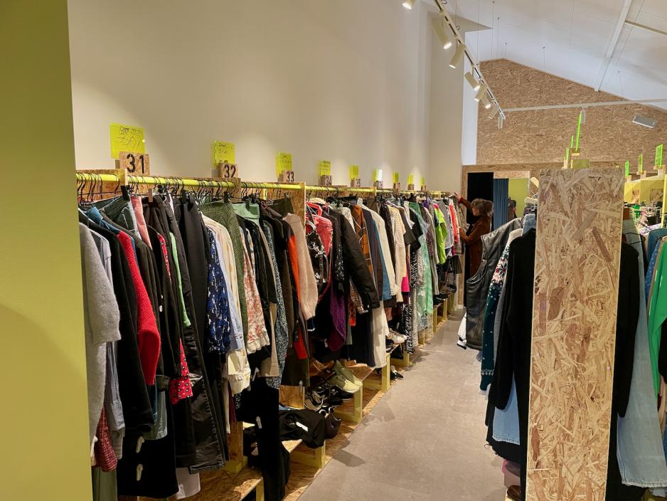 Kl&aelig;r p&aring; stativ i et stort lyst rom. P&aring; enden av stativene ser en dame p&aring; kl&aelig;r.