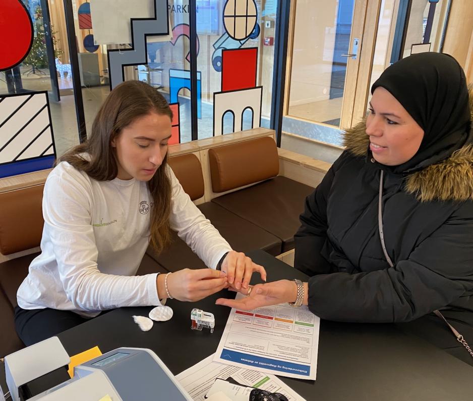 Kvinne f&aring;r m&aring;lt blodsukker. Hun smiler til kvinnelig frisklivsveileder, som holder konsentrert p&aring; med m&aring;lingen. 