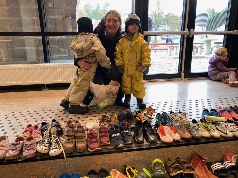 En dame med to barn, en p&aring; hver side av seg, sitter p&aring; huk foran mange sko som kan byttes. 