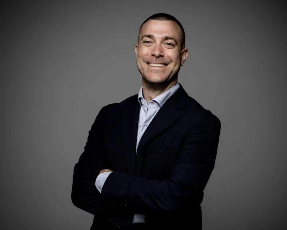 Portrett av Jan Thomas Holmlund fra Dagbladet. Han st&aring;r foran en gr&aring; bakgrunn, og har p&aring; seg svart blazer og bl&aring; skjorte. Han har armene i kors, og smiler inn i kamera.