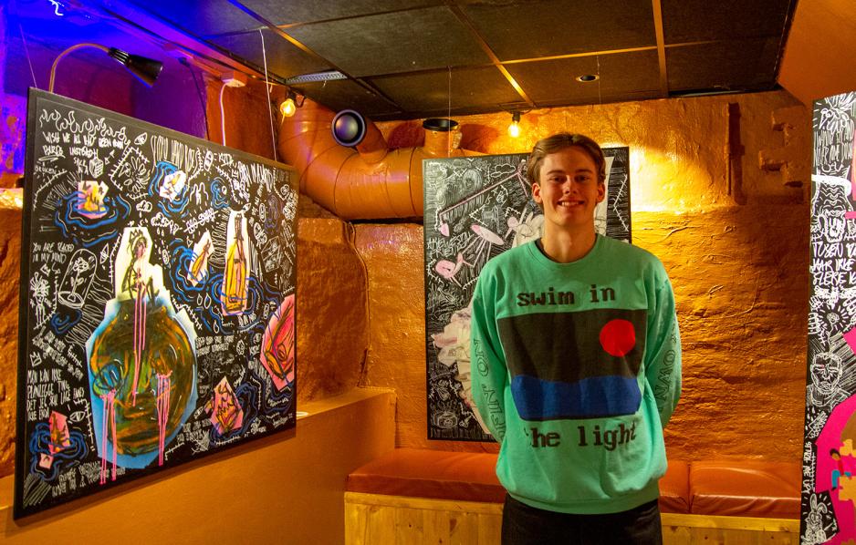 En ung gutt smiler til kamera. Han har p&aring; en turkis genser med et motiv. Han st&aring;r foran flere utstilte bilder. 