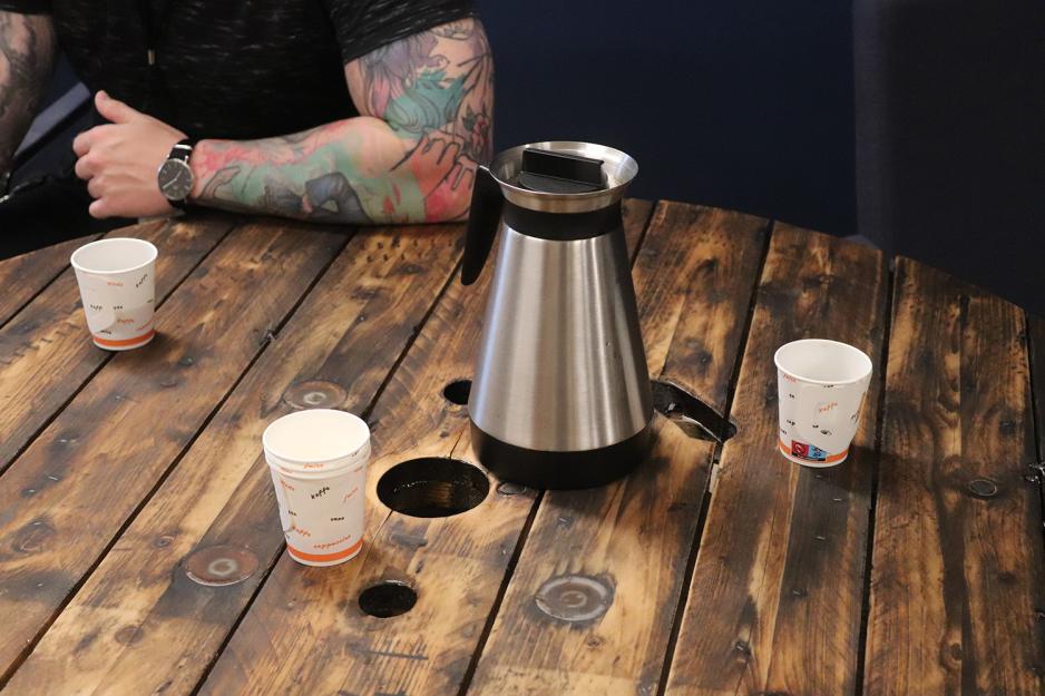 En kaffekanne og tre pappkopper p&aring; et trebord. En tatovert arm lener seg p&aring; bordet.