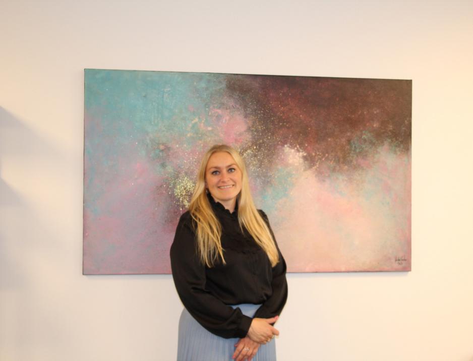 Blond kvinne st&aring;r ved maleri med rosa, bl&aring; og gull farger. 