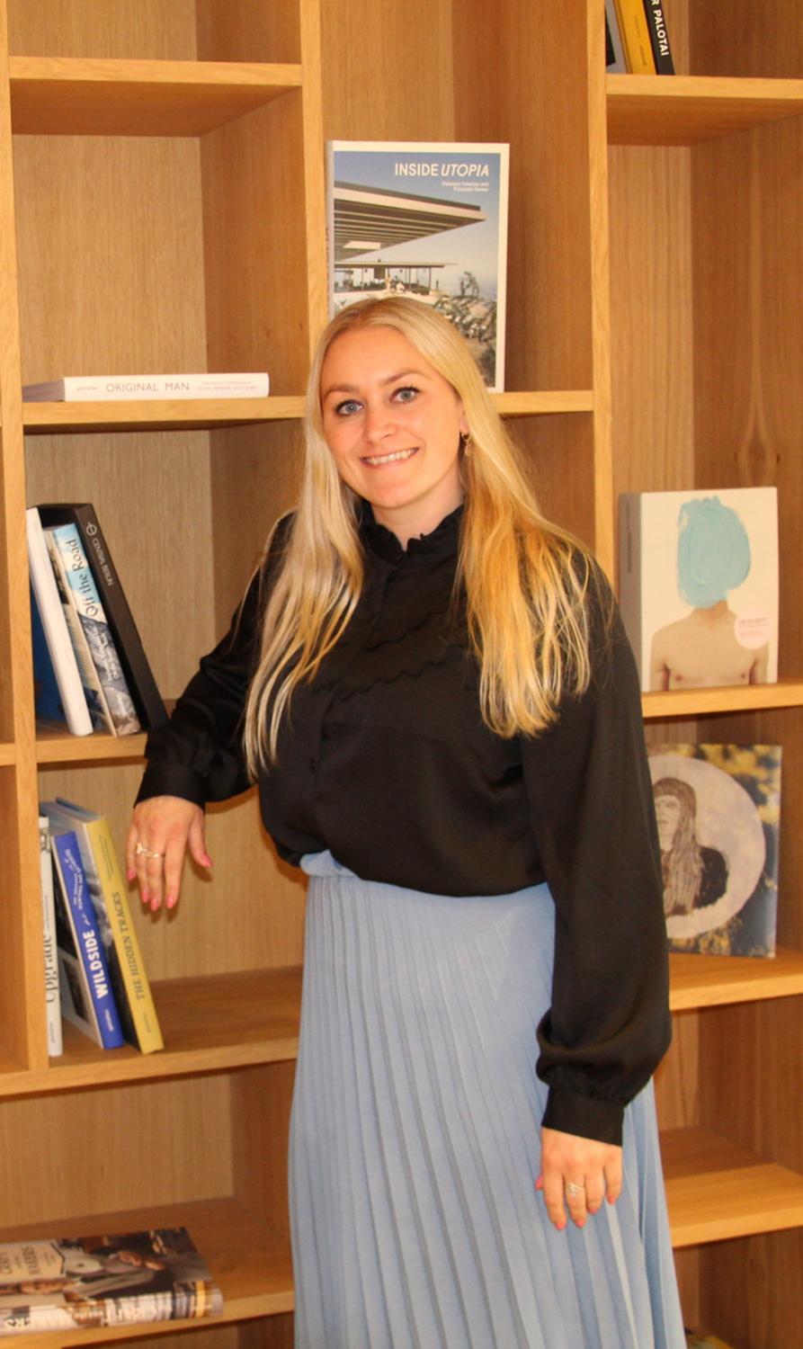 Blond kvinne st&aring;r ved bokhyllen. 