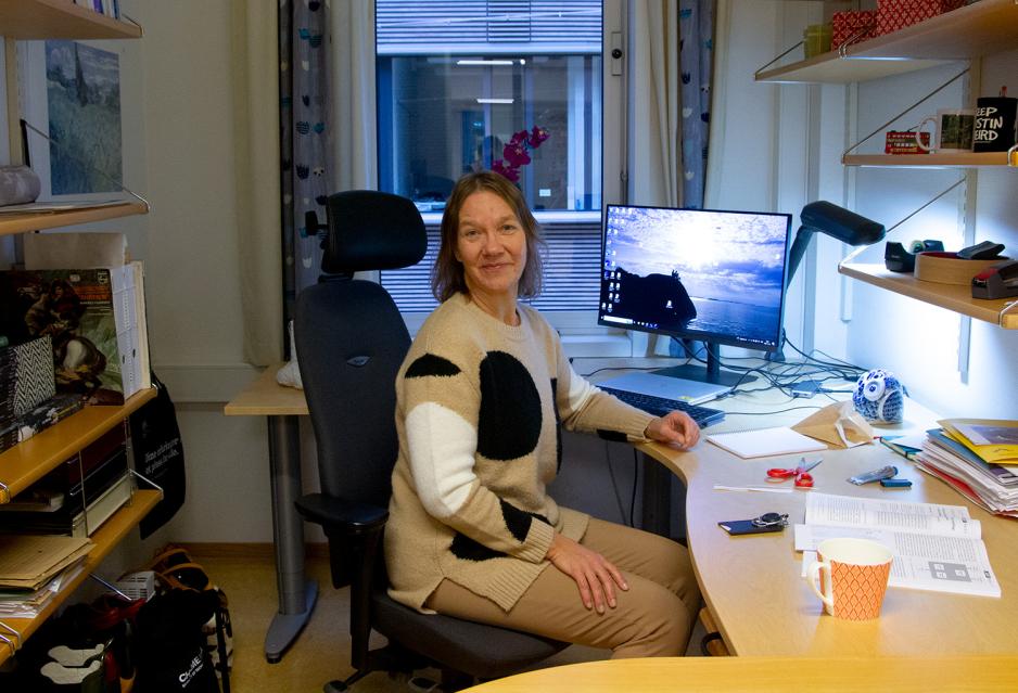 En dame sitter smilende p&aring; kontoret sitt. Hun har brukt, skulderlangt h&aring;r, med to store PC-skjermer bak seg. 