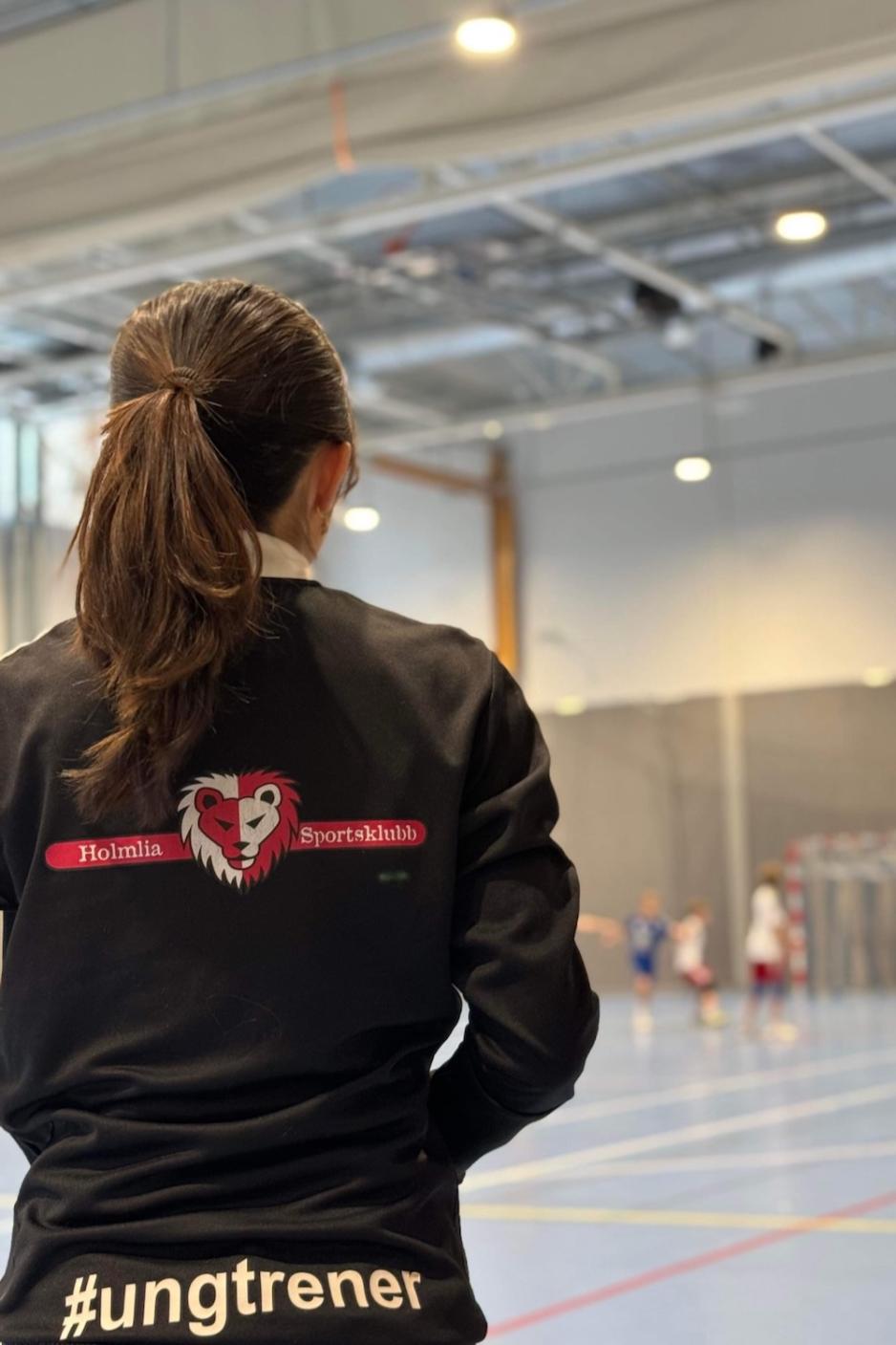 Katrina Lu med Holmlia sportsklubb jakke med ung trener p&aring; ryggen i h&aring;ndballhallen
