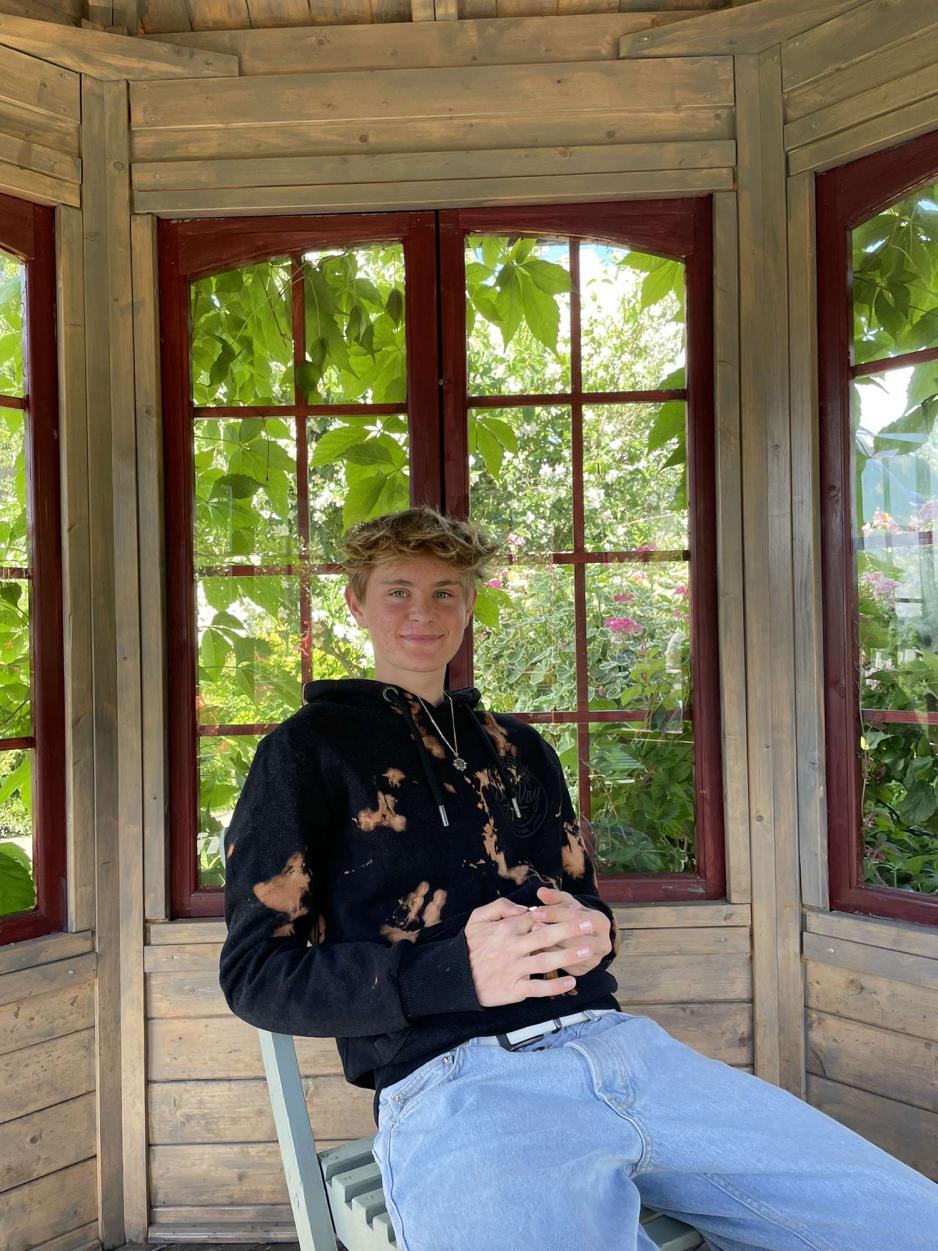 BIlde av en blond gutt med bl&aring; &oslash;yne som sitter i en stol. Gutten har p&aring; en svart hettegenser med brune flekker og lysebl&aring;e bukser. Han har armene i fanget og et kjede rundt halsen. bak han er det et vinduet som er dekket til av gr&oslash;nne planter fra utsiden.