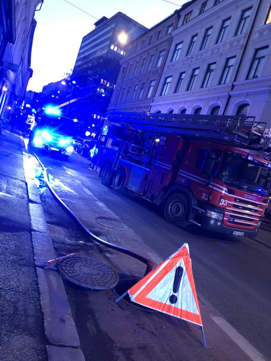 En vannledning ligger bortover langs veien, den kommer opp fra et &aring;pent kumlokk med en varseltrekant foran. I bakgrunnen st&aring;r brannbiler og ambulanse og bl&aring;lys lyser kaotisk utover - situasjonen/f&oslash;lelsen p&aring; stedet er litt uoversiktlig.