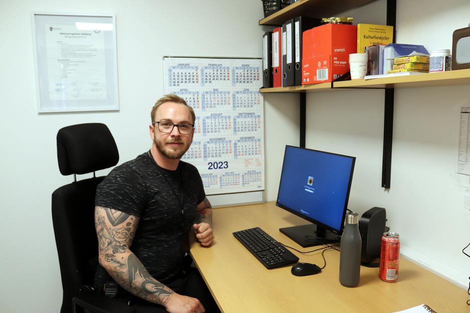 En mann sitter p&aring; et kontor. Rundt p&aring; veggene er det forskjellige plakater. Det er en hylle over bordet. 