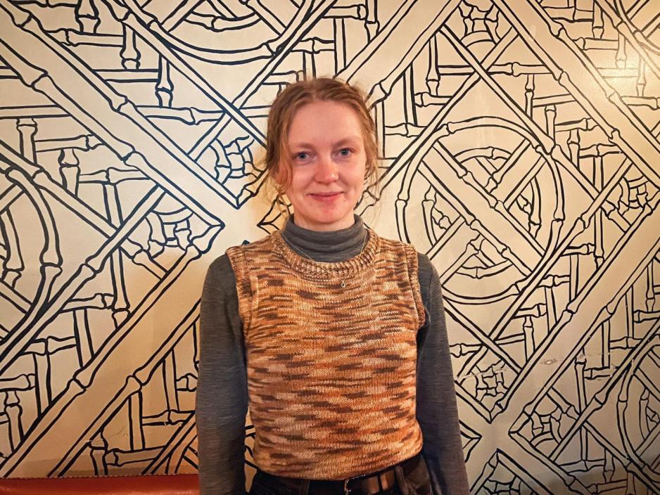 Ei blond smilende dame foran en vegg som er svart og hvit. Hun har p&aring; seg  en langhals genser og en vest over. 